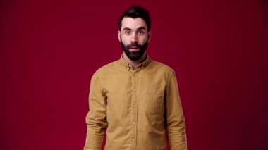 Başardım, şok oldum, çok mutluyum, gülümsüyorum, adam kamerayı işaret ediyor, siyah saçlı, sakallı, kahverengi gömlek giyiyor, arka plan kırmızı