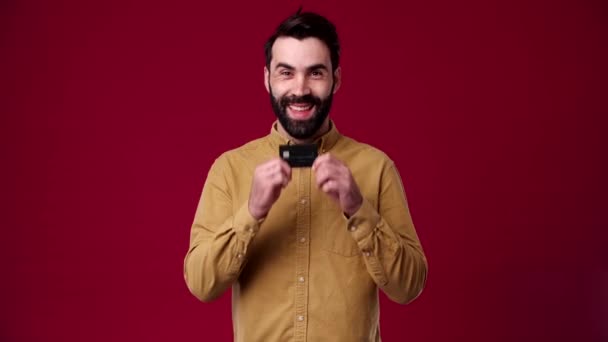 男は手に銀行カードを持っていて 彼はそれが好き 彼は笑顔 茶色のシャツに身を包んだ 背景が赤 — ストック動画