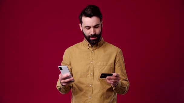 男はオンラインで買い物をしてる銀行カードと携帯電話を手に持ってる黒髪で髭を生やして茶色のシャツを着てる — ストック動画