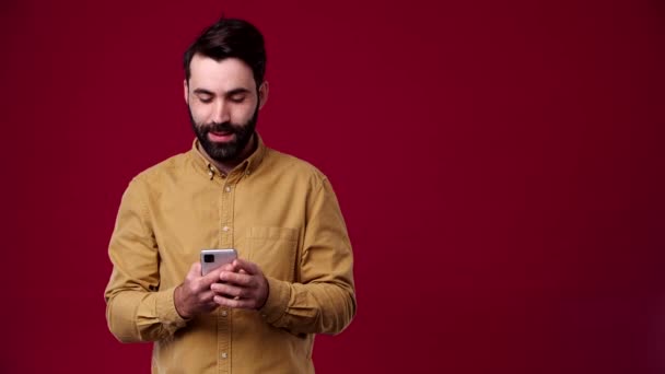 这个人手里拿着手机 指着旁边 空旷的空间 他笑着 穿着一件棕色的衬衫 背景是红色 — 图库视频影像