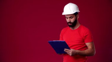 Bir çalışan dosyaya veri yazıyor, koruyucu bir kask takıyor, dosya mavi, sakallı, koyu renk saçlı, kırmızı tişört giyiyor, arka plan kırmızı.