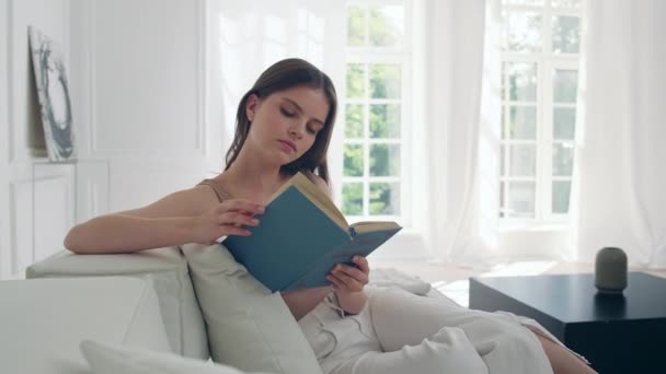 Chica joven en la sala de estar en el sofá está descansando leyendo un libro. — Vídeos de Stock