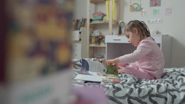 Маленькая девочка читает книгу на кровати в детской.. — стоковое видео