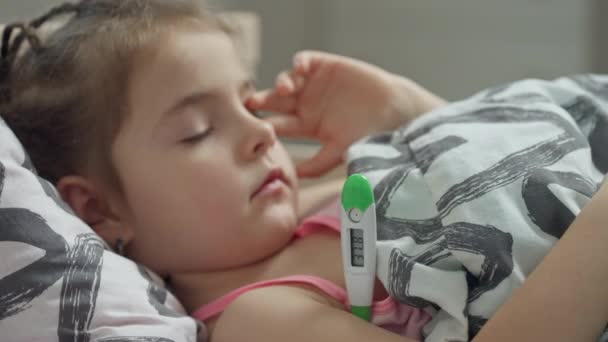Маленькая больная девочка лежит в постели с термометром, который измеряет температуру — стоковое видео