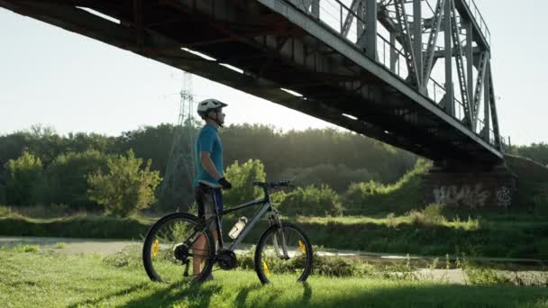 Парень ездит на велосипеде по траве в красивом пейзаже с мостом — стоковое видео
