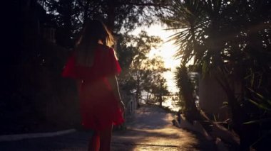 Kırmızı elbiseli kız deniz kenarında palmiye ağaçlarıyla sokakta yürüyor..