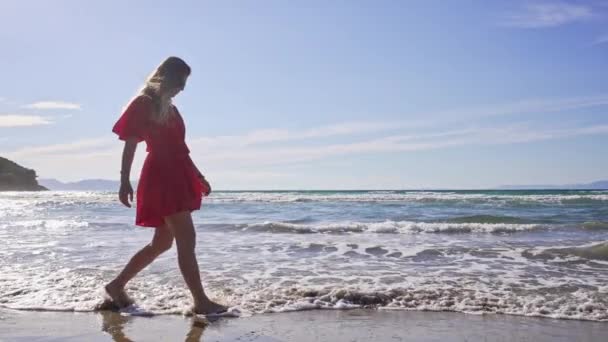 Une fille de race caucasienne en robe rouge se promène sur une plage de sable fin au bord de la mer. — Video
