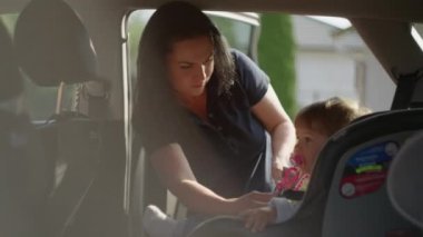 Anne kızını çocuk oto koltuğuna bağlar..