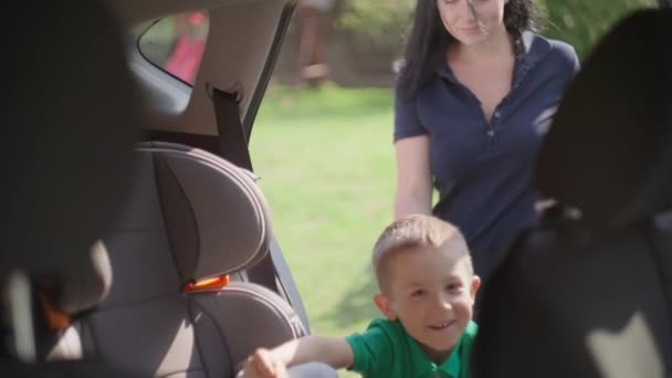 Η μαμά και ο γιος πάνε στο αυτοκίνητο, το παιδί μπαίνει σε παιδικό κάθισμα.. — Αρχείο Βίντεο