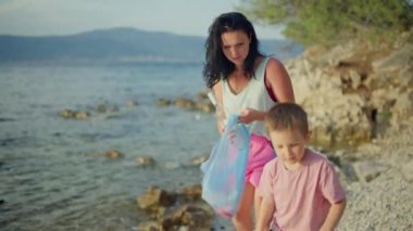 Anne ve küçük oğlu sahili temizliyor. Çöplerle dolu güzel bir manzara ve aile çöp torbasında plastik ve diğer atıkları toplayarak temizliyor..