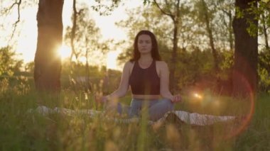 Doğayla uyum içinde olan kız gün batımında yoga yapıyor. Park 'ta, Asana' yı çimlerin üzerinde meditasyon yaparken gösteren yaşam tarzı videosu..