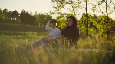 Anne, oğluyla birlikte parkta çimlerin üzerinde oturuyor. Anne bebekle oynuyor, mutlu aile manzarada gülümsüyor..