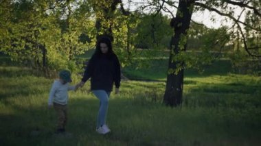Anne ve oğlu parkta yürüyüş yapıyor. Ağaçlar, çimenler ve güneşte yürüyen bir kadından bir çocuk, takım çalışması ve ormanda aile tatili..