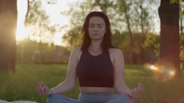 Parkta meditasyon yapan bir kız. Yoga yapan bir kadın ağaçların ve güneş ışınlarının yanındaki çimlerin üzerinde oturuyor. İnsan doğayla ve kendisiyle baş başa..
