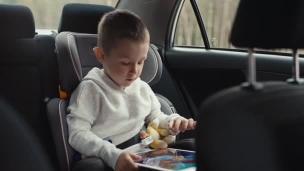 男の子は車で旅行中にタブレットでゲームをプレイします 子供は子供の車の座席に座ってガジェットを演奏する時間を過ごす — ストック動画