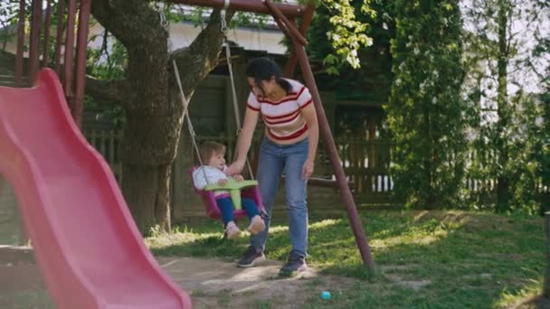母と娘は遊び場に住んでいます 美しい風景の中でスイングに子供に乗る女性 チームワークと幸せな子供時代 — ストック動画
