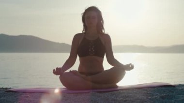 Kadın gün batımında pencerenin yanında kollarını kavuşturarak meditasyon yapıyor. Kız tek başına yoga ve spor yapıyor. Sağlıklı bir yaşam tarzı..