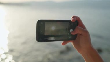 Telefonun kadın ellerinde yakın plan çekimi. Gün batımında deniz kenarında fotoğraf çekiyorlar. Hayatın mutlu anları.