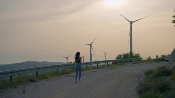 風車の近くでジョギングしている女の子 夕日の美しい山の風景の中でアクティブなライフスタイルスポーツや環境エネルギーの源 — ストック動画