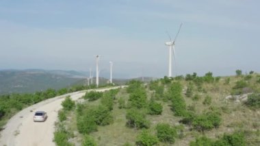 Yel değirmenlerinin yakınındaki yolda giden hava manzaralı araba. Ekolojik elektrik üretimi ve seyahat eden insanlar.