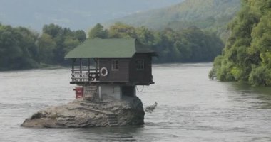 Sırbistan 'da Drina nehrindeki bir kayanın üzerinde duran dağ, ağaç ve manzarayla çevrili bir evin güzel videosu.