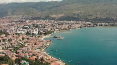 Ohri Gölü kıyılarında kırmızı çatıları olan evleri ve dağlarla çevrili tekneleri olan iskeleleri olan antik bir kentin insansız hava aracı videosu. Ohri Makedonya 'nın havadan görüşü.