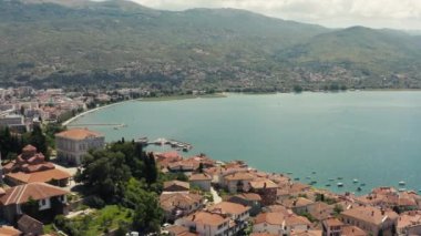 Ohri Gölü kıyılarında kırmızı çatıları olan evleri ve dağlarla çevrili tekneleri olan iskeleleri olan antik bir kentin insansız hava aracı videosu. Ohri Makedonya 'nın havadan görüşü.