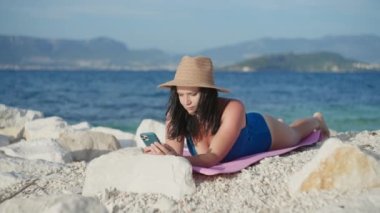 Woman sunbathes on the beach and writes on the phone. Girl orders food and makes purchases online from a gadget by the ocean in a swimsuit.