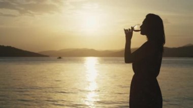 Okyanusun kenarında gün doğumunda şarap içen bir kız. Denizde tatilde elinde bir bardak alkol olan bir kadının silueti..