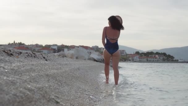 岩の多いビーチを歩くダンジョンでの女性のスローモーションビデオ 美しい風景の中で海で休暇中の女の子のライフスタイルビデオ — ストック動画
