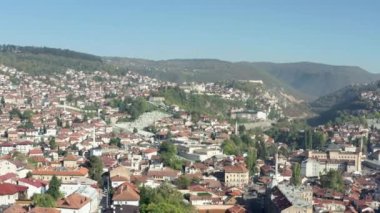 Bosna-Hersek 'in havadan görüşü. İnsansız hava aracı, dağların ve gökyüzünün manzarasında duran kırmızı çatıları olan evlerin üzerinde uçuyor. Dağ sıraları ve zirveler görülebilir durumda..
