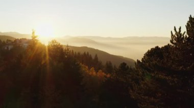 Gün batımının insansız hava aracı videosu, dağlarda gün doğumu ön planda ağaçlarla kaplı sıradağları görebilirsiniz, yamaçlar sisle kaplıdır ve güneş ışınlarıyla kaplıdır. Havadan manzara görüntüsü.