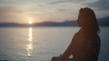 Close-up portrait of a girl by the sea at sunset. Beautiful woman on a alone sitting by the ocean and looking at the waves and the sun.
