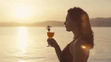 Mayo giymiş bir kızın gün batımında deniz kenarında kokteyl içerken çekilmiş yakın çekim videosu. Güzel manzara ve yaz tatilinde okyanus kenarında dinlenen bir kadın. Yüksek kalite 4k görüntü