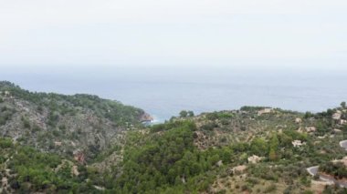 Houses in the mountains by the ocean dron 4K video. Aerial view of mountain ranges with buildings, forests with trees that stand on the shoreline of the sea where the waves crash against the rocks.
