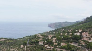 Okyanus dron 4K videosunun yanındaki dağlarda evler. Dağ manzarası binalarla, dalgaların kayalara çarptığı deniz kıyısındaki ağaçlarla çevrilidir..