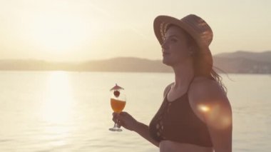 Slow motion video of a girl with a cocktail in her hands walking on the beach at sunset. Smiling and happy woman by the ocean resting. High quality 4k footage