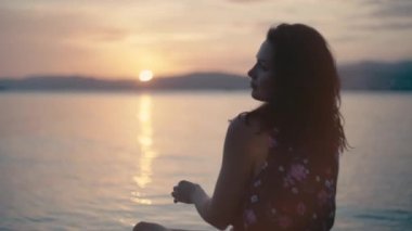 Romantic Caucasian girl in a dress by the sea. Sunset ocean waves and pensive beautiful woman looking at the sun relaxing in beautiful scenery.