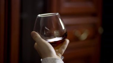 Bir adamın elinde bir bardak konyak. Adamın biri elinde brendi bardağını çeviriyor. Evinde alkolle rahatlayan bir adamın 4K videosu. Yüksek kalite 4k görüntü