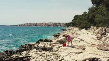 Hava manzaralı kız deniz kenarındaki bir kayanın üzerinde güneşlenmeye geldi. Yaz tatilindeki kadın, okyanus yakınlarındaki güzel bir manzarada mayo giymiş..