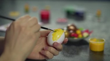 Paskalya için hazırlanıyorum. Dişi el, fırçayla sarı bir Paskalya yumurtası boyuyor. Bir tatil yumurtasının yavaş çekim 4K videosu. Yüksek kalite 4k görüntü
