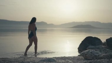 Slow motion video of a beautiful Caucasian woman entering the ocean at sunrise. woman goes swimming in the sea at sunset in the mountain scenery.