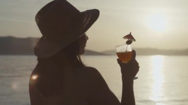 Slow motion video of a girl drinking a cocktail at sunset by the sea. Happy girl on vacation by the ocean relaxes and rejoices.