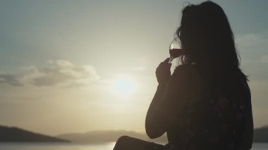 Gün batımında deniz manzarasında şarap içen bir kız. Romantik bir buluşmada okyanusun kenarında oturan ve elinde bardakla gün batımını izleyen bir kadın..