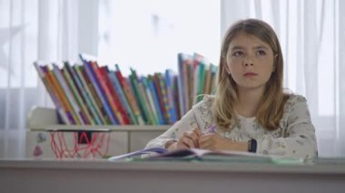 The girl thinks and then writes in a notebook. Homework preparation. Teenager training at home. The child sits at a desk and writes.