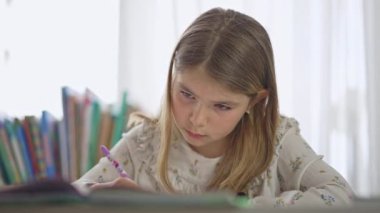 European girl learn home lesson. Kid write school lesson at home. Young girl look book and write after.