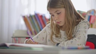 Schoolgirl writes in a notebook at the table. Portrait of a child teaching homework. Teenager prepares lessons for school in the home room with books on the background.