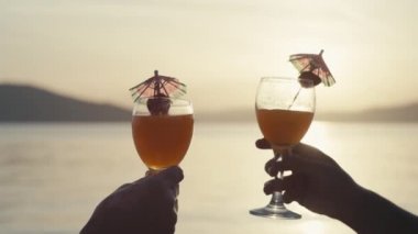Slow motion close-up of knocking glasses with cocktails at sunset. Beautiful seascape and people relaxing and drinking drinks by the ocean.