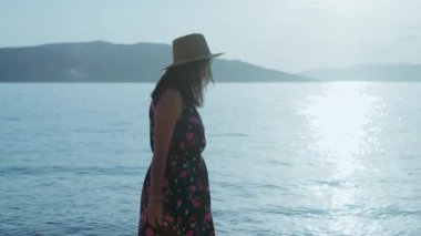 A carnivorous girl in a hat and dress walks on the seashore. Relax and rest by the ocean at sunset. High quality 4k footage
