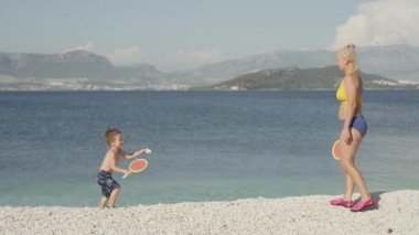 Çocuk ve annesi sahilde tenis oynuyorlar. Aktif aile tatili, bir çocuk ve raketli bir kadının yavaş çekim videosu, mutlu ve gülümseyen. Yüksek kalite 4k görüntü